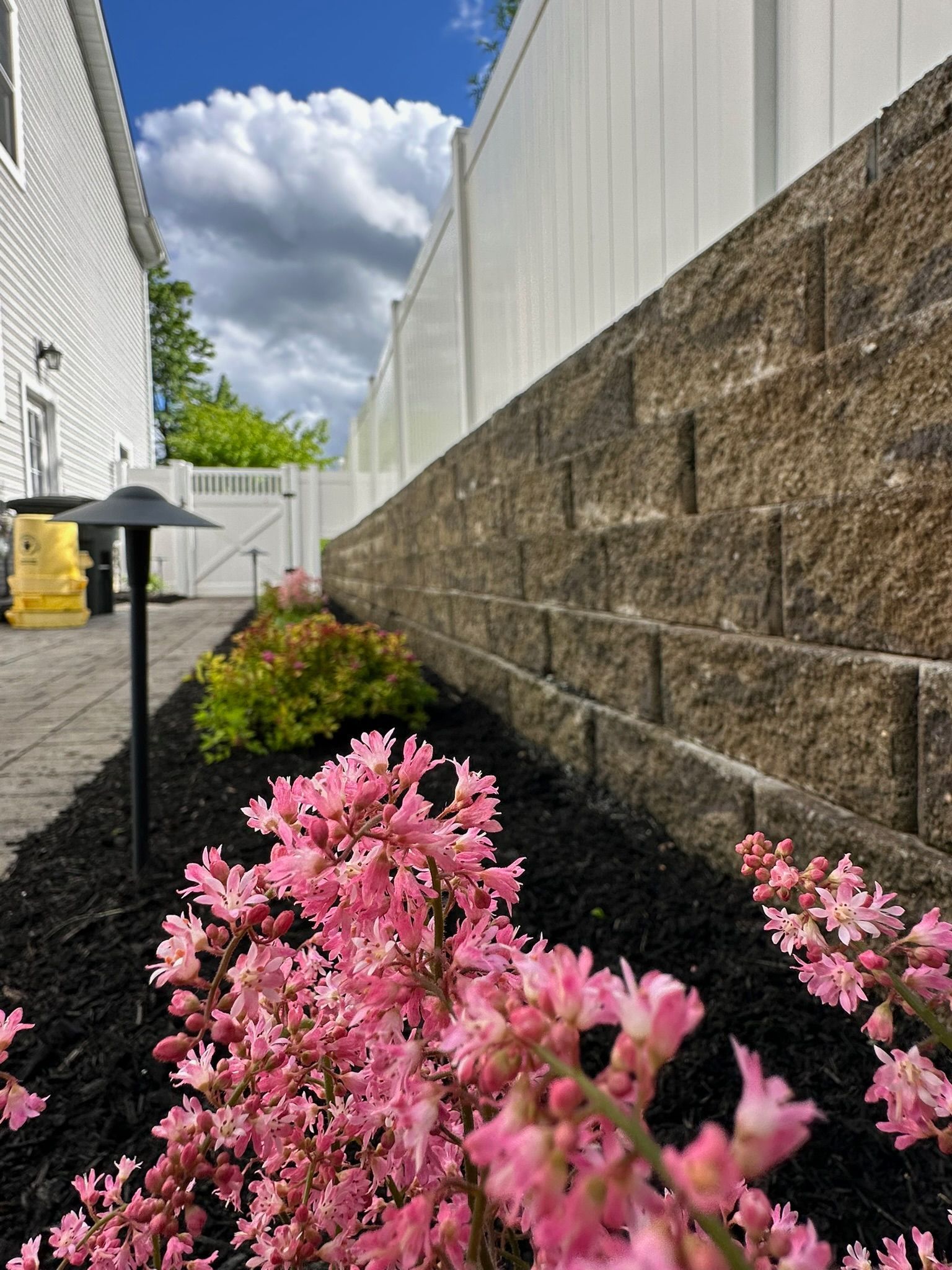 Pink flowers in front of a brick retaining wall. Black mulch, white fence, and cloudy sky.