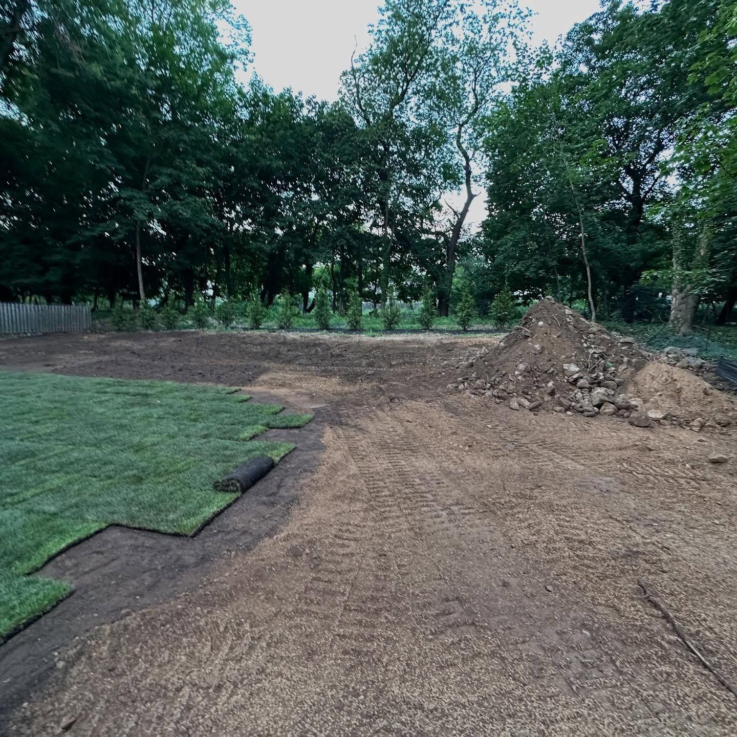 Laying sod in a yard; fresh green grass contrasts with prepared soil and a pile of dirt, trees in the background.