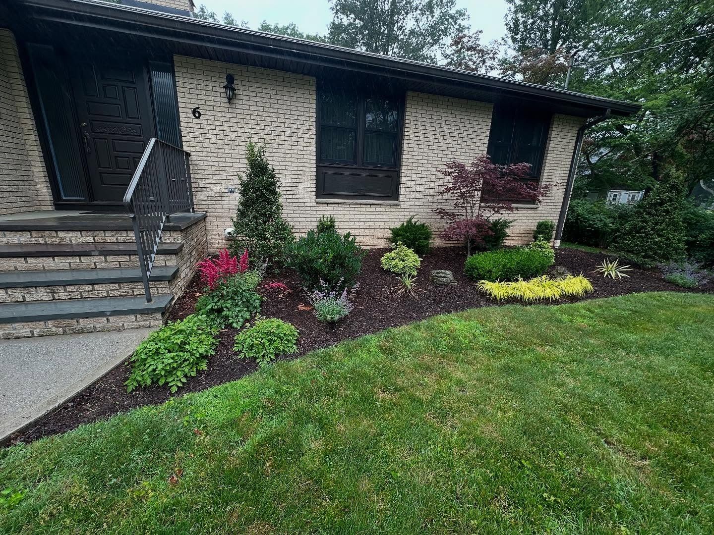 A landscaped front yard with a brick house, colorful plants, and dark mulch.