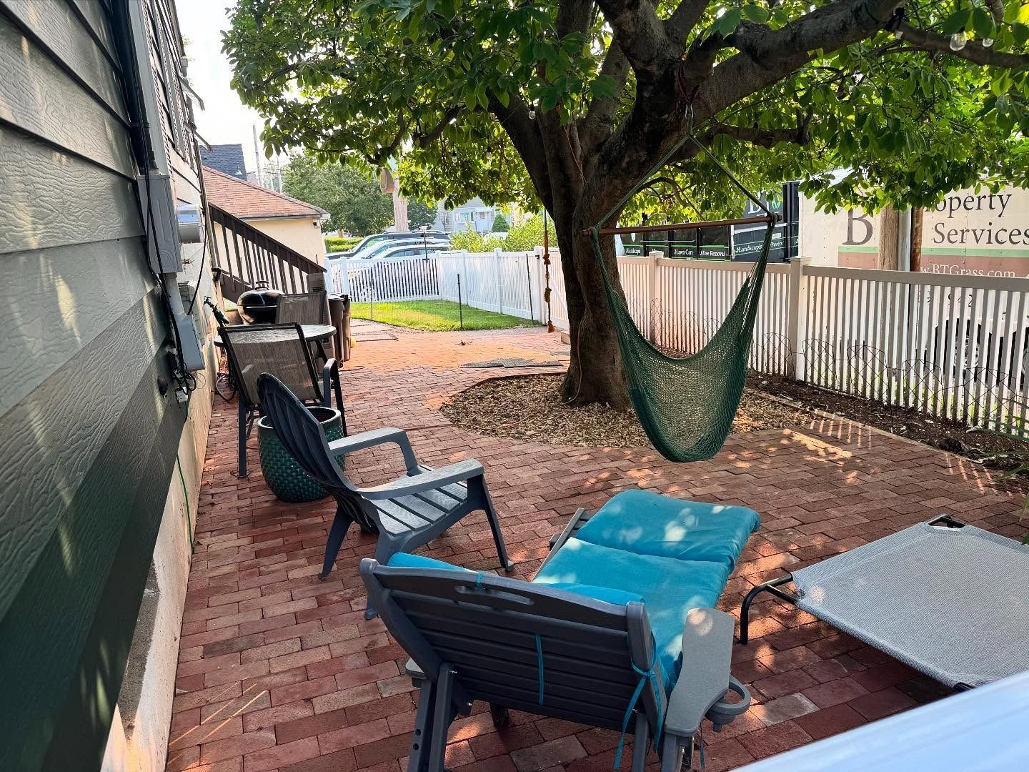 Backyard patio with brick, chairs, and a hammock under a tree, near a white picket fence.