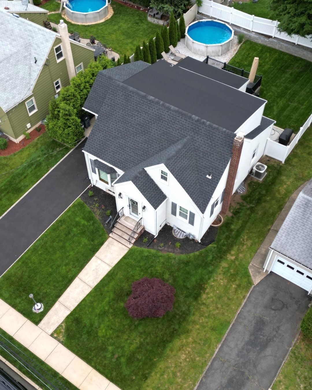 Aerial view of a white house with a black roof, green lawn, and a driveway. Two pools in the background.