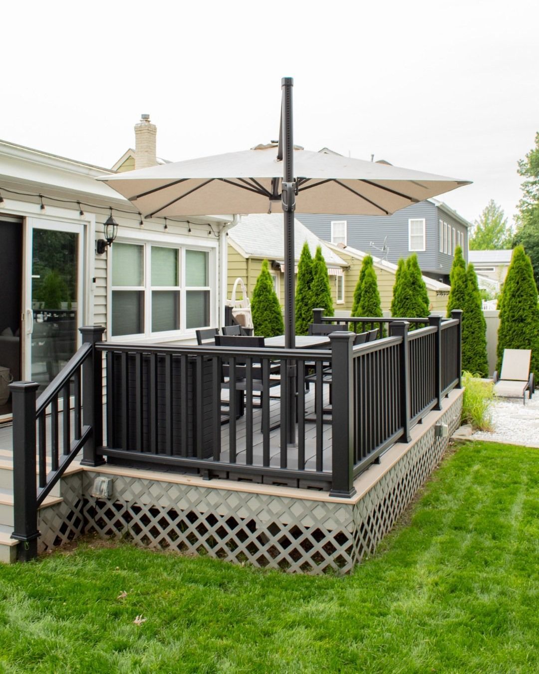 Black deck with beige umbrella, table, and chairs. Green lawn and house in the background.