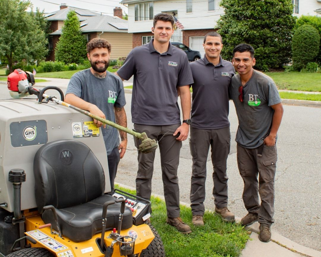 Four landscapers pose by a riding mower and string trimmer on a residential street.