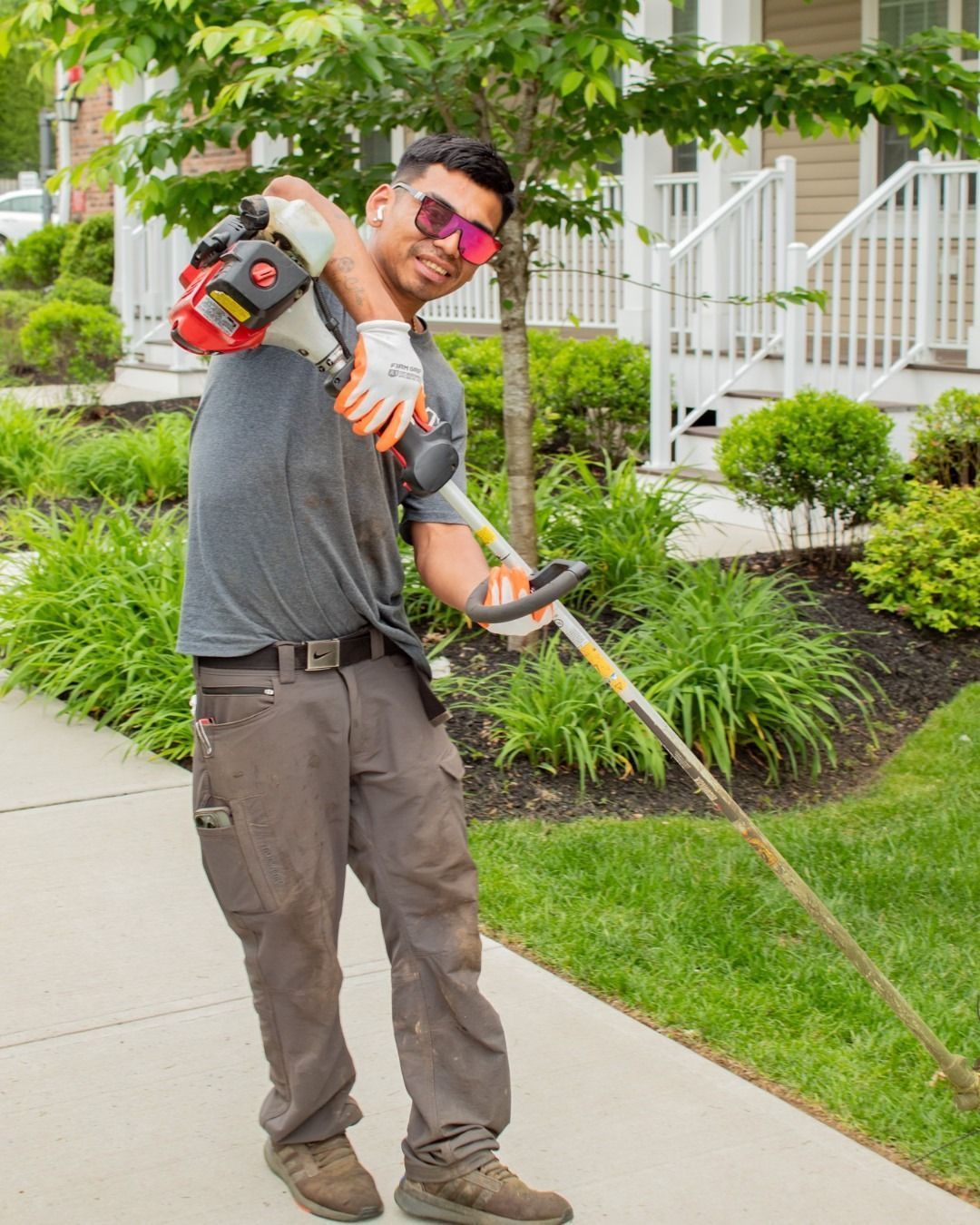 Man with a weed whacker on a sidewalk, wearing sunglasses, smiling, and gardening near buildings.