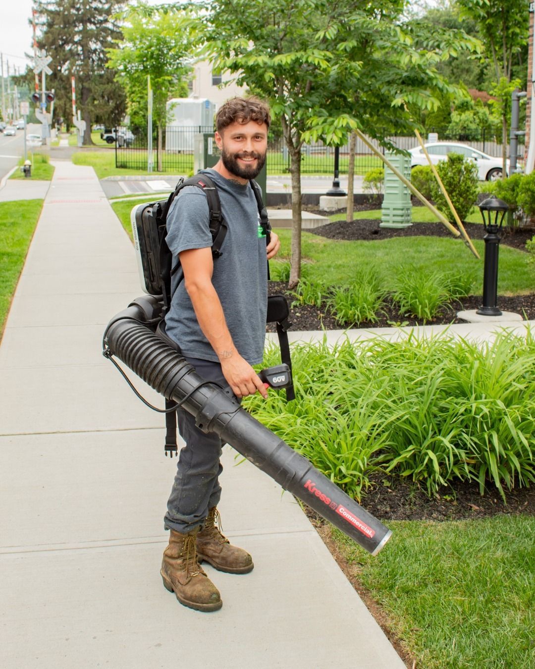 Man with a backpack leaf blower, standing on a sidewalk next to a lawn, smiling.