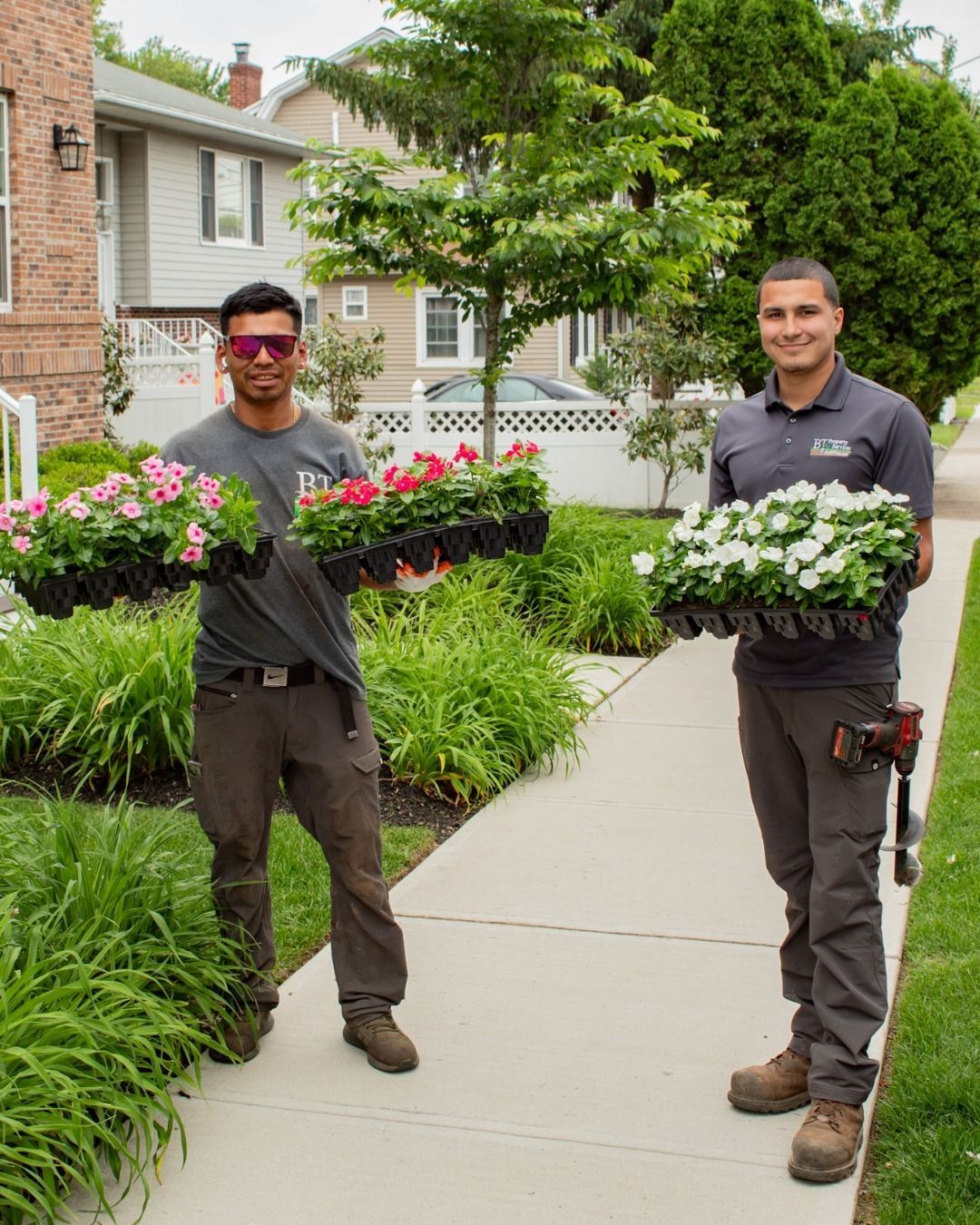 Two men in work clothes holding trays of colorful flowers, standing on a sidewalk near a building.