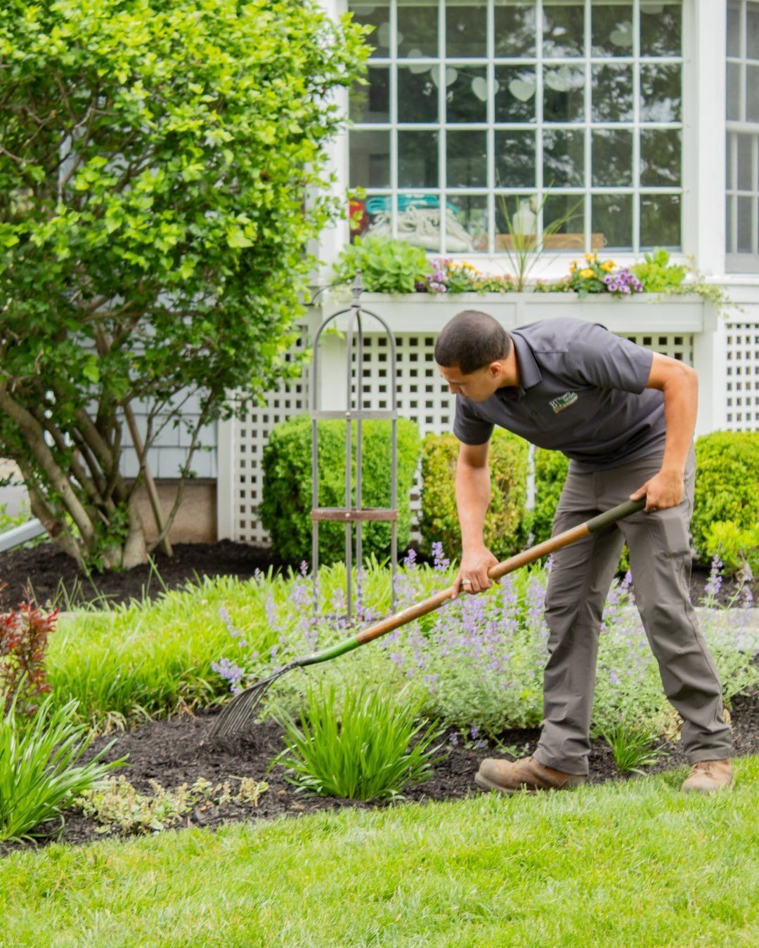 Man rakes soil in a flower bed in front of a white house.