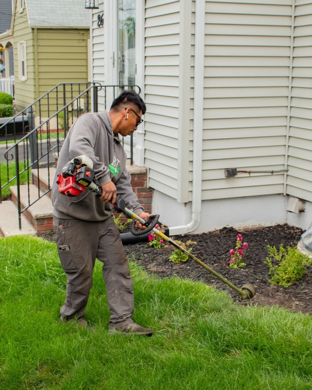 Man in grey hoodie trimming grass with a weed whacker near a house.