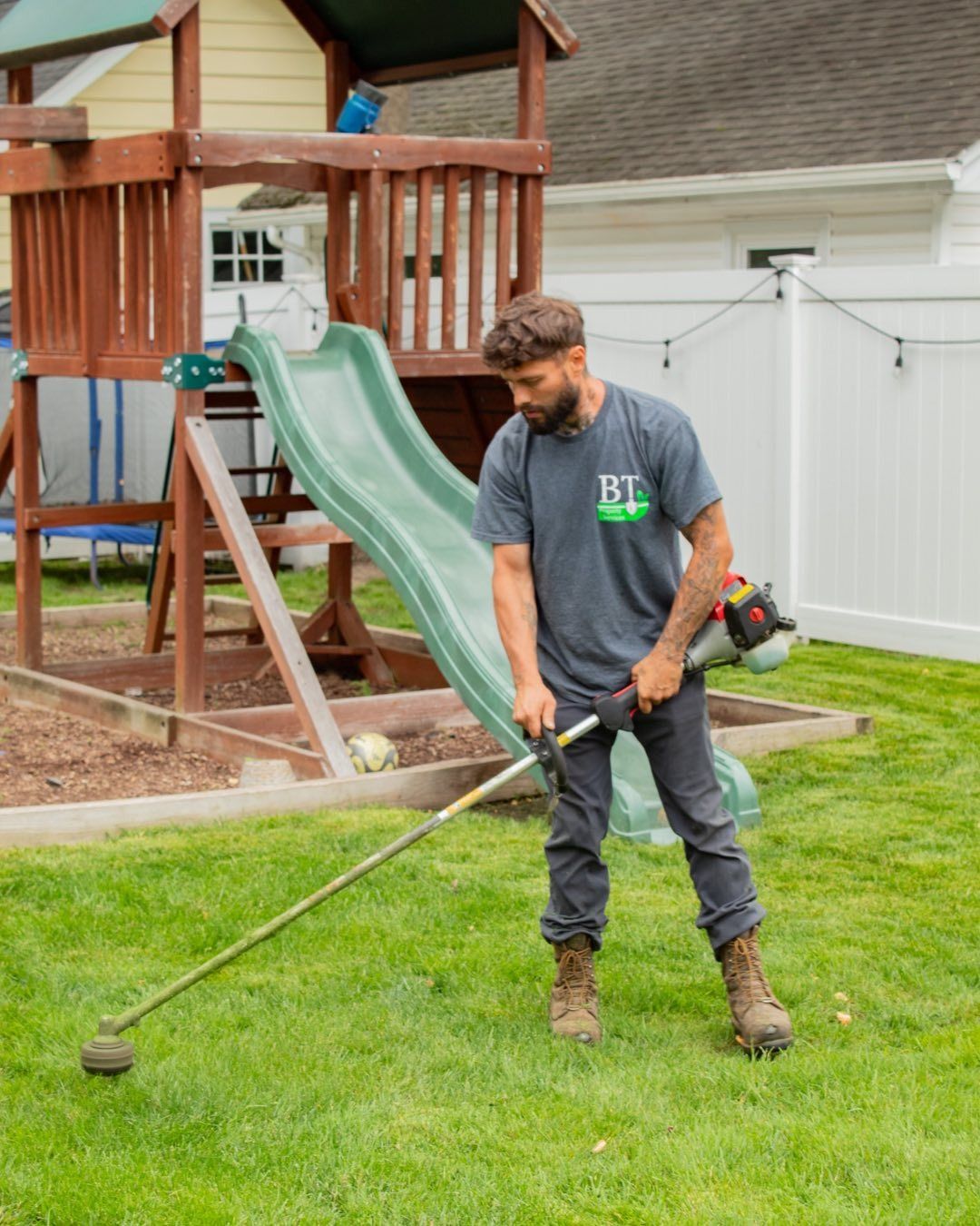 Man using a weed wacker in a backyard near a wooden playset.