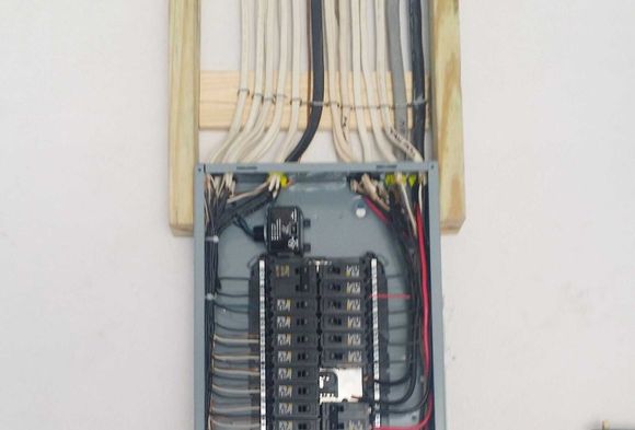 Electrical panel with wires and circuit breakers mounted on a white wall.