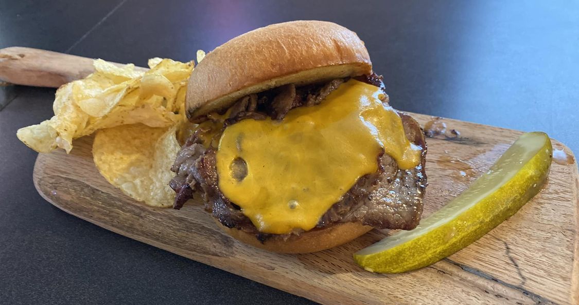 A hamburger with cheese, potato chips and a pickle on a wooden cutting board