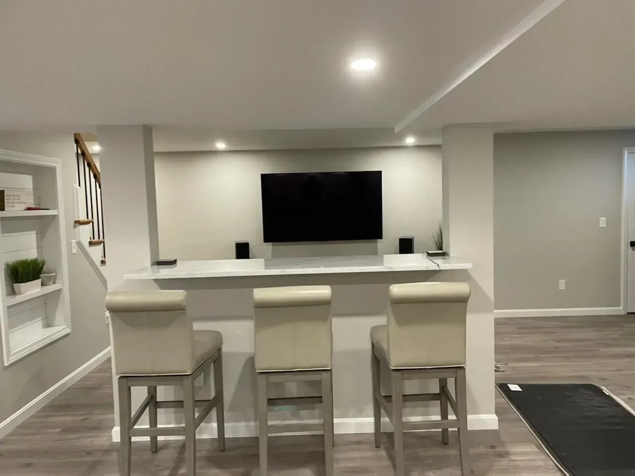 Modern open-plan basement with white bar counter, three stools, wall-mounted TV, and recessed lighting