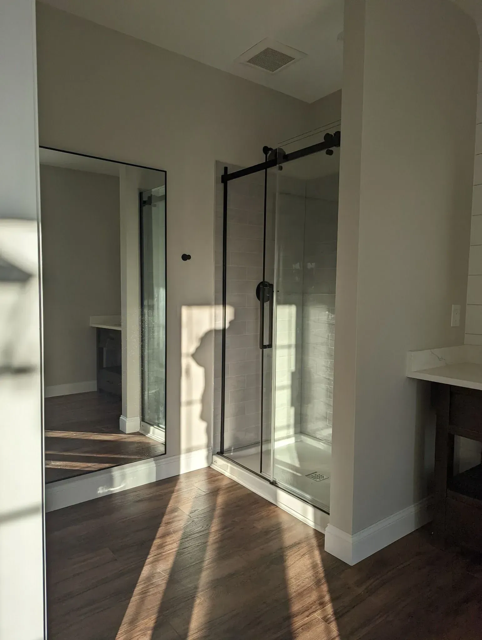 Empty modern bathroom corner with glass shower enclosure and mirrored wall, sunlight on hardwood floor
