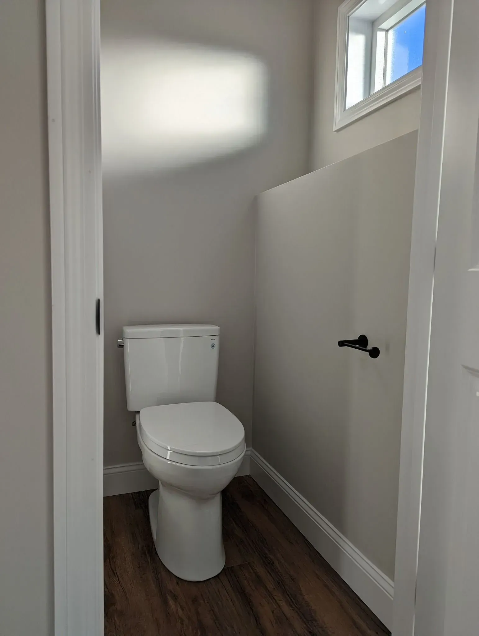 Small bathroom with a toilet, wood floor, white walls, and a high window.