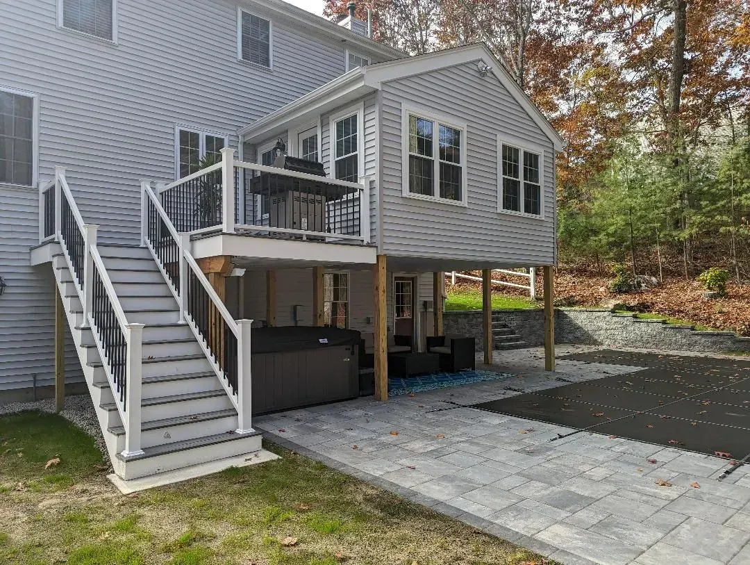 Raised gray deck with stairs, covered patio, and paved walkway beside a house in a wooded yard