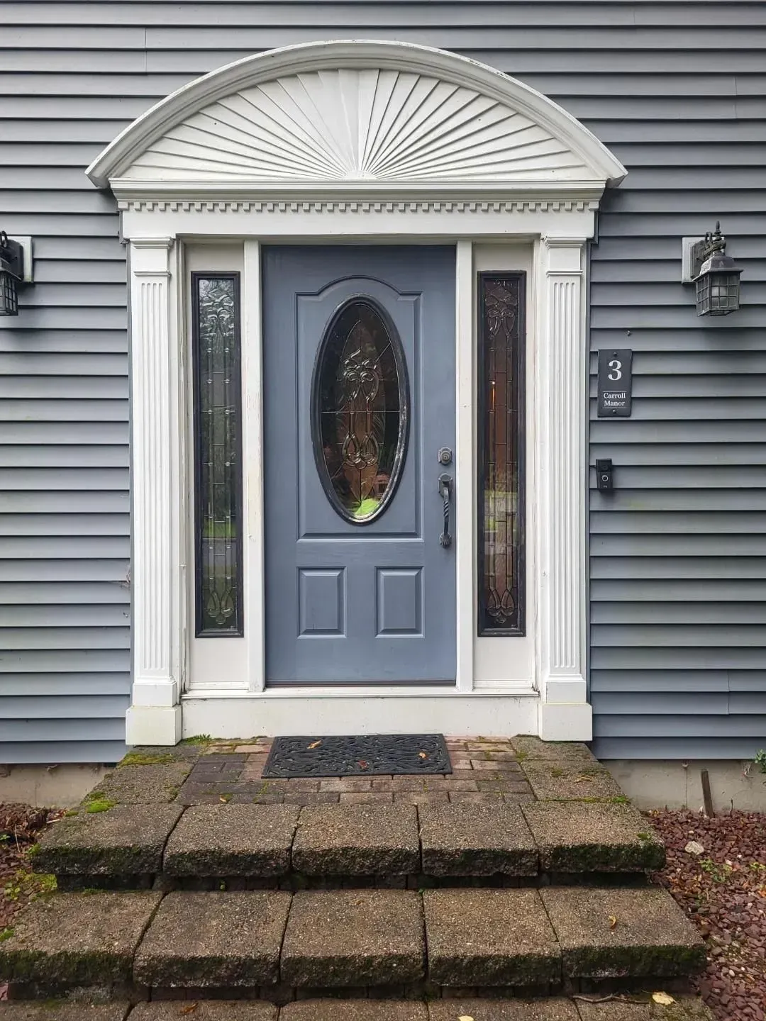 Blue front door with oval glass, framed by white trim and small stone steps in front of a gray house.