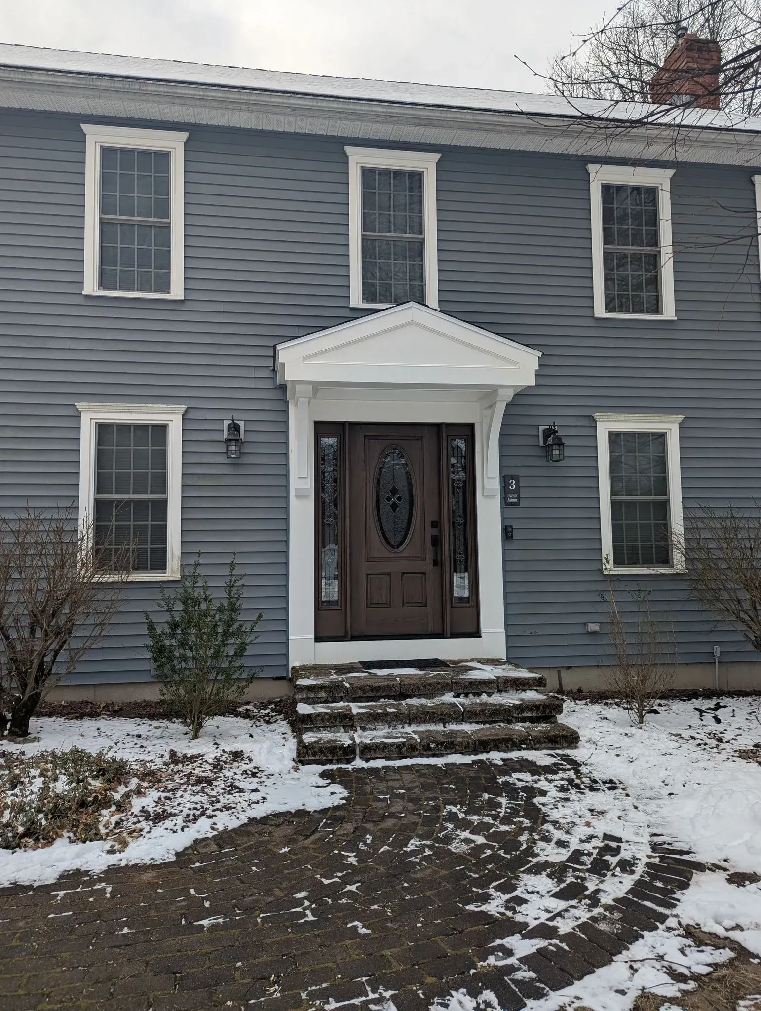 Blue two-story house with white trim, front steps, and a brown door, with snow on the ground.