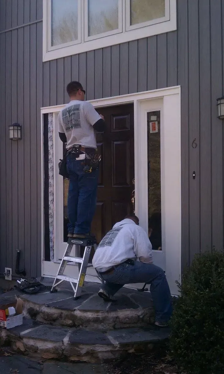 Two workers installing a door on a gray house, one on a ladder and one crouching below.