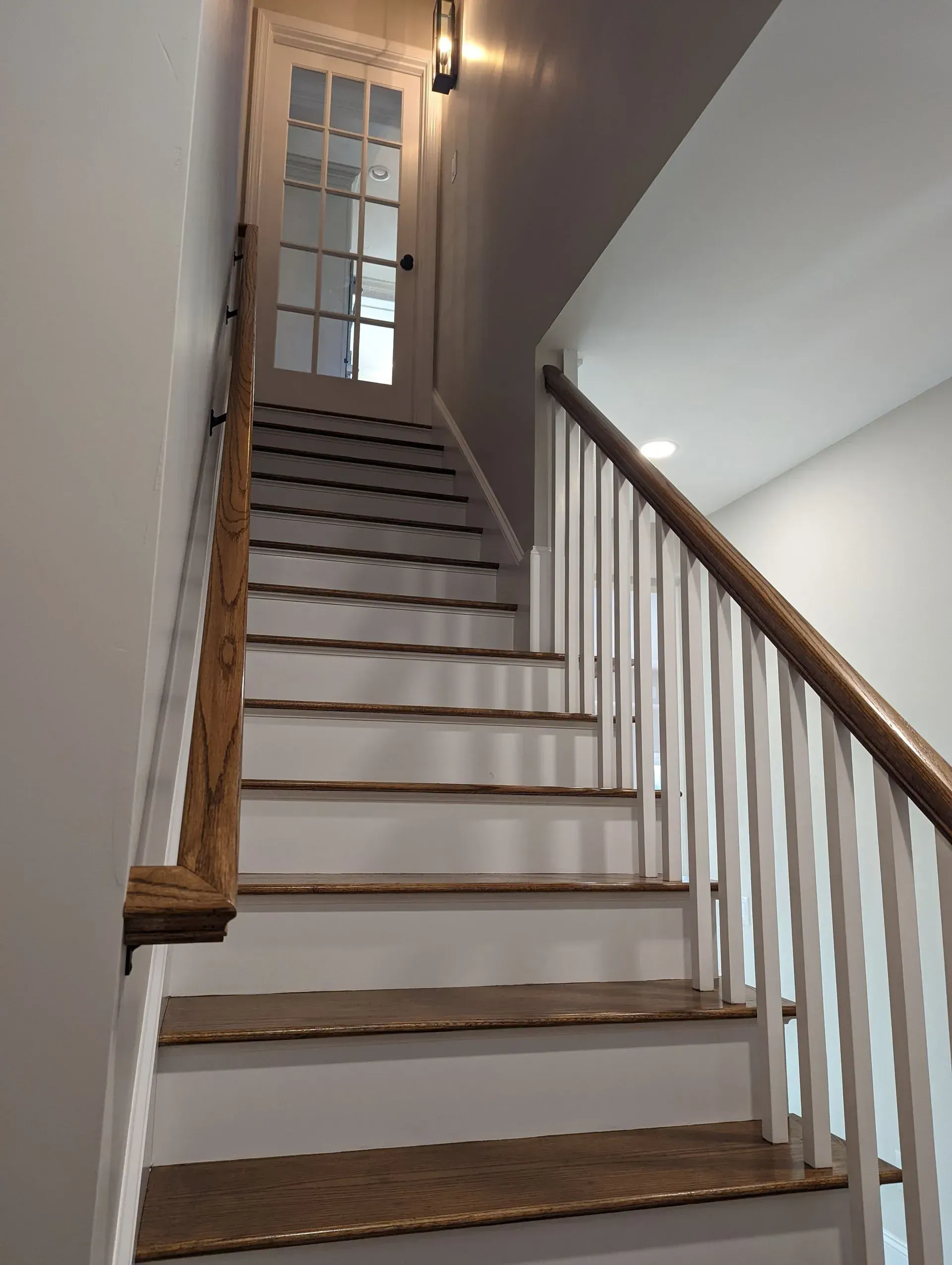 Narrow indoor staircase with wooden handrail, white walls, and a door at the top.