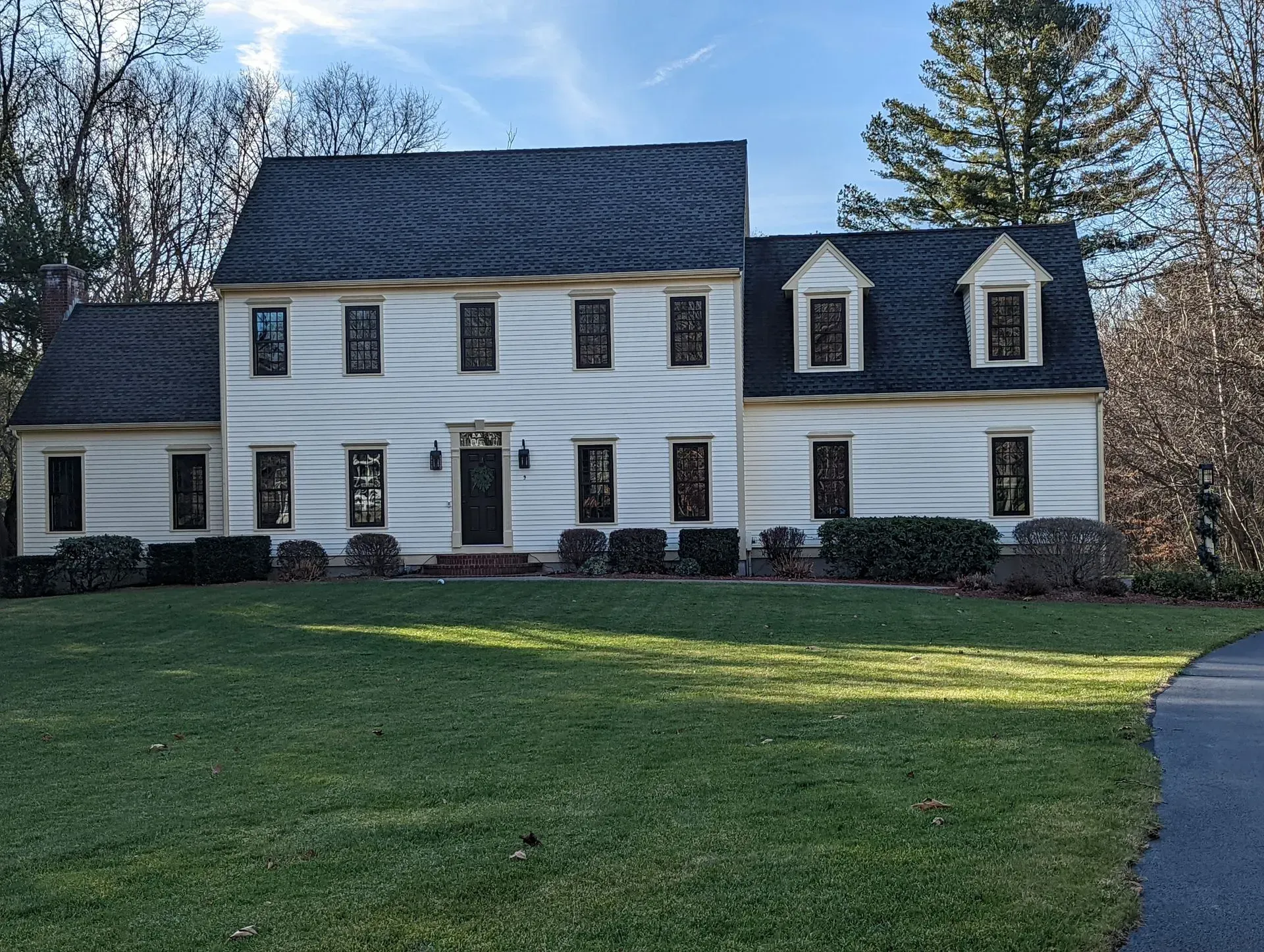 White colonial-style house with dark roof and dormer windows on a large green lawn under a blue sky