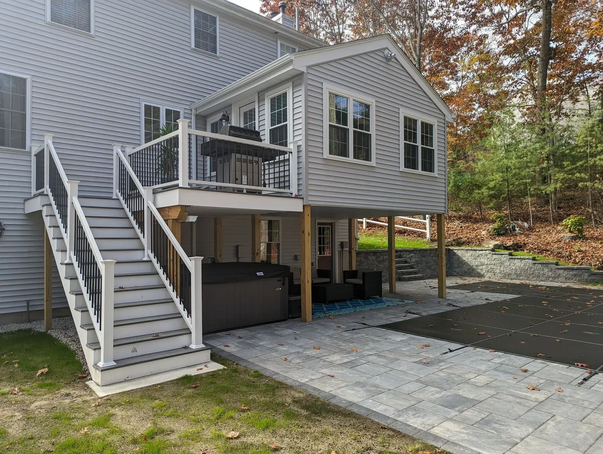 Backyard deck with stairs, railing, and storage beneath a raised gray house beside a paved patio