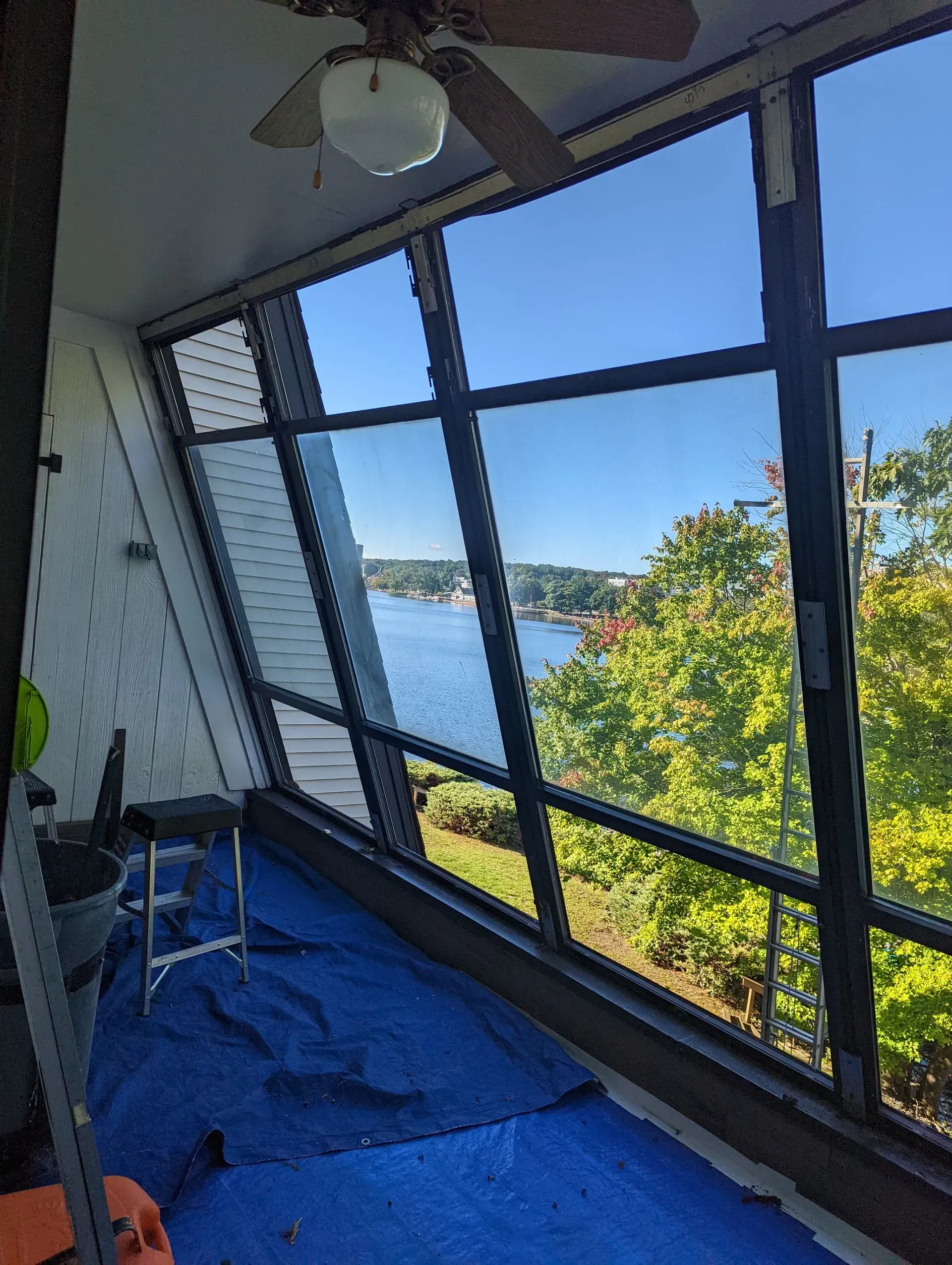 Enclosed balcony with slanted windows, blue floor covering, and a river view beyond green trees.