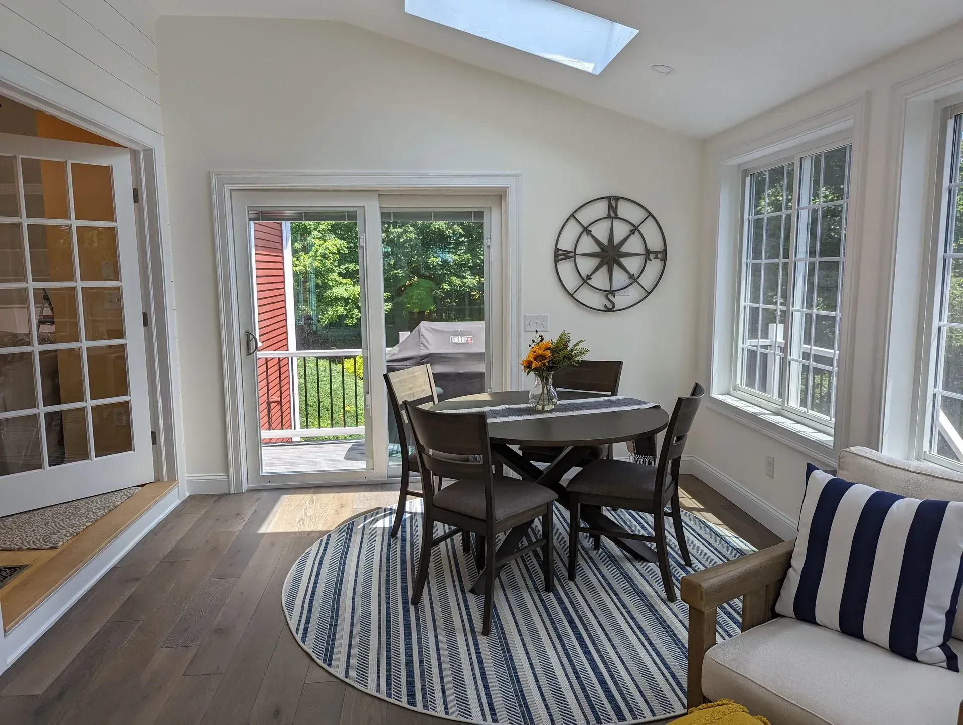 Bright sunroom dining area with round table, striped rug, wicker chair, wall clock, and open doors to a deck