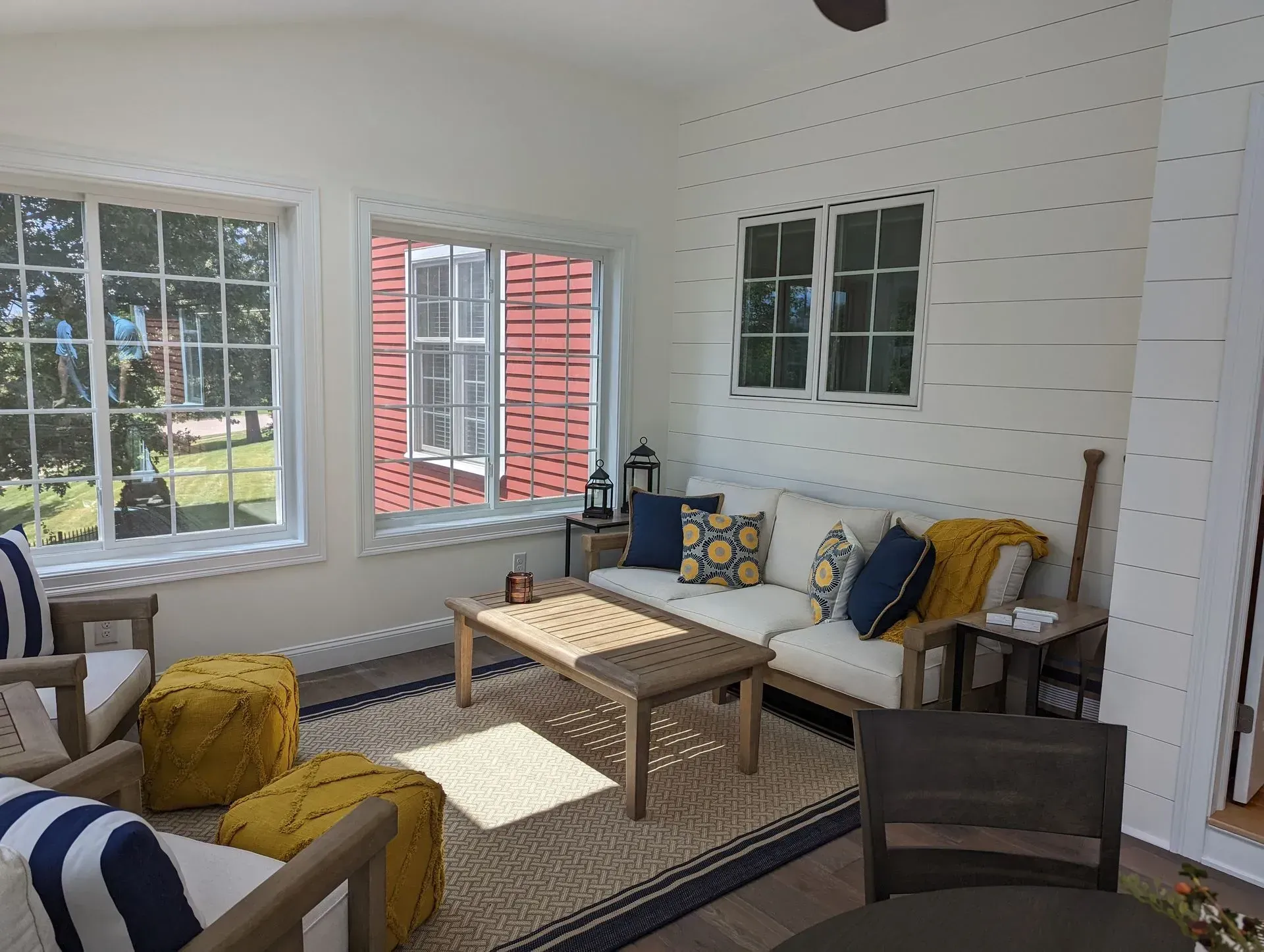 Bright sunroom with white shiplap walls, a sofa, chairs, rug, and wooden coffee table by large windows.
