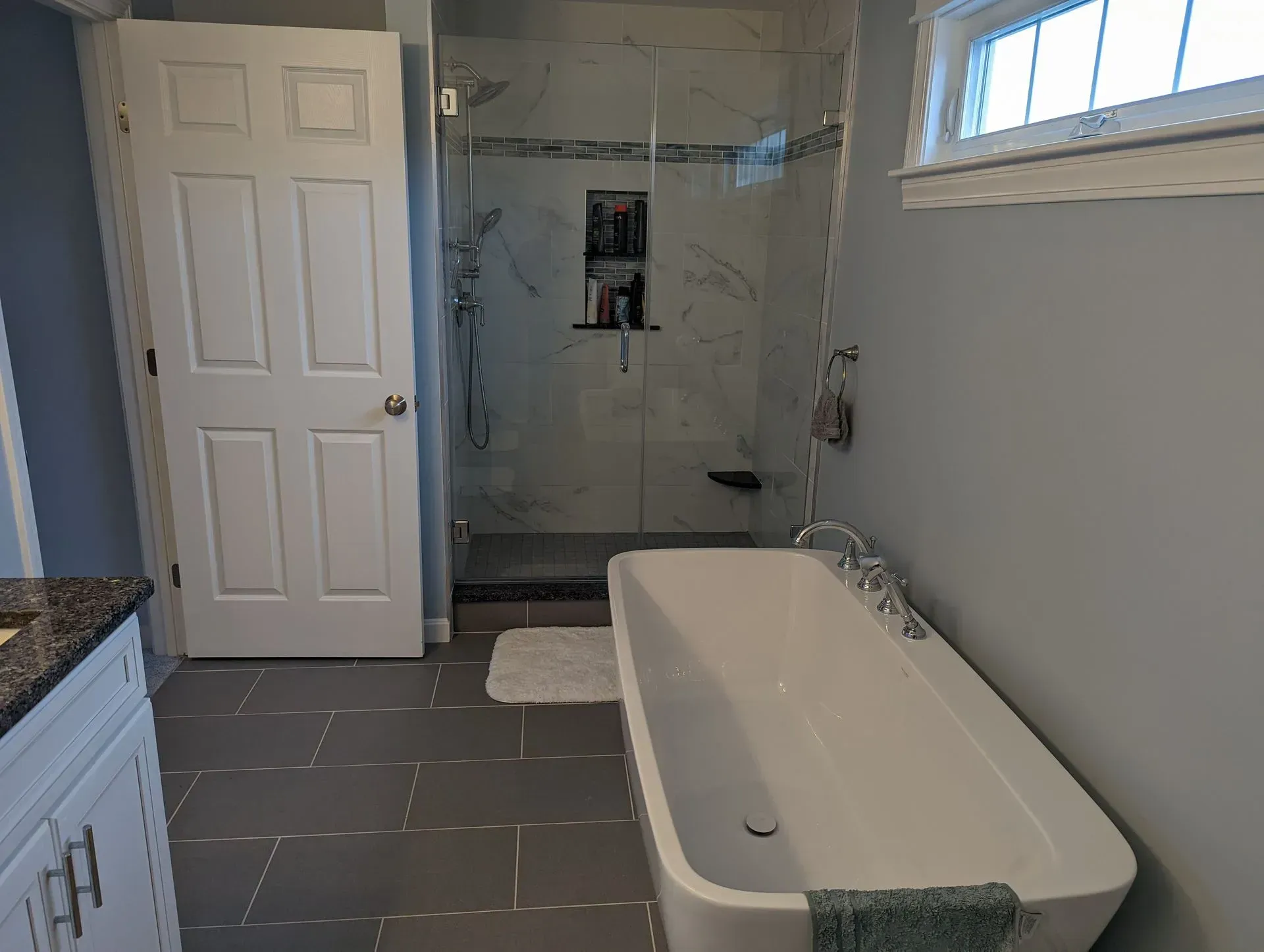 Bathroom with freestanding tub, tiled floor, shower alcove, and white cabinets, viewed from the doorway.
