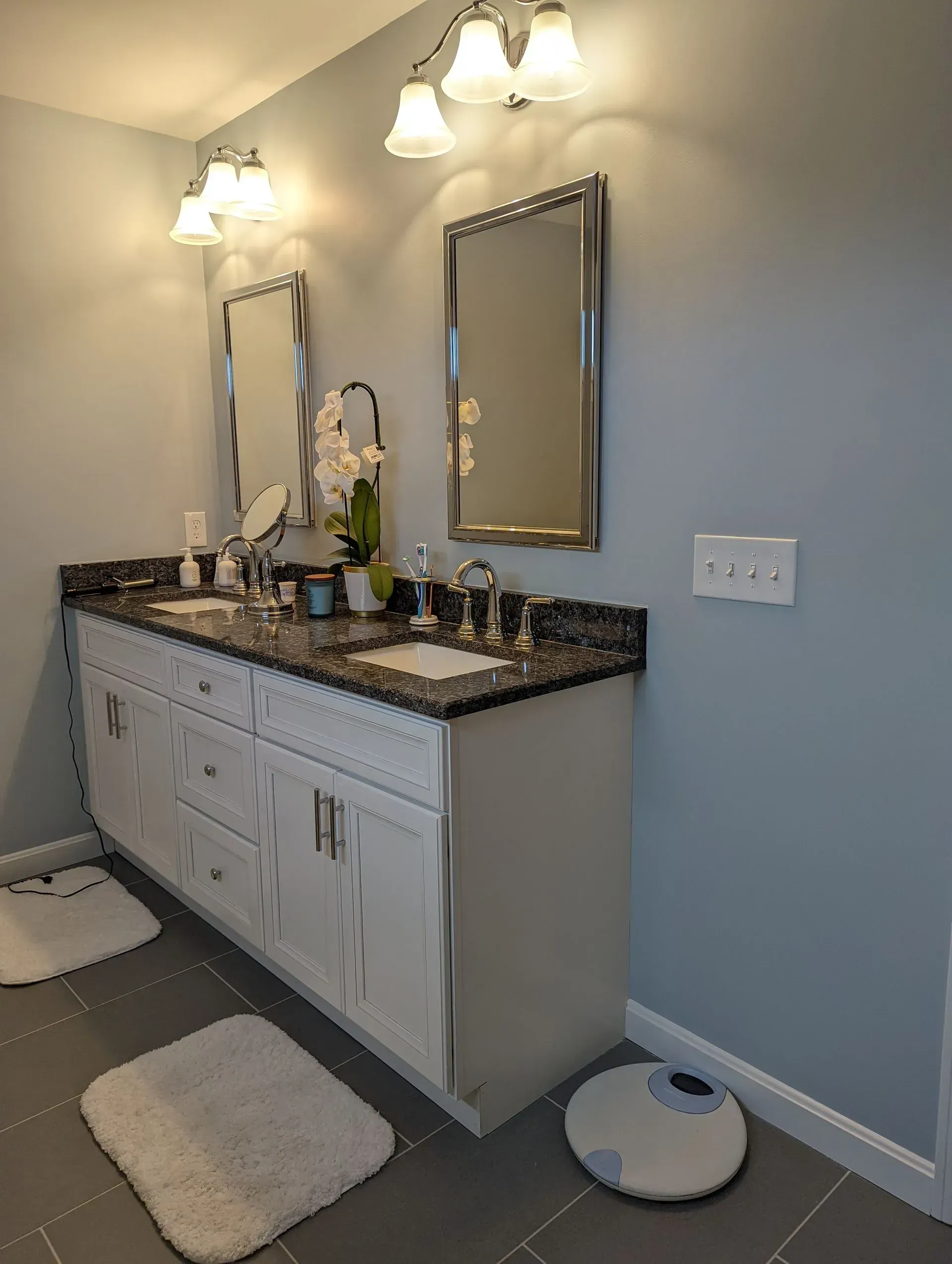 Bathroom vanity with double mirrors, granite countertop, white cabinets, and a robot vacuum on the floor