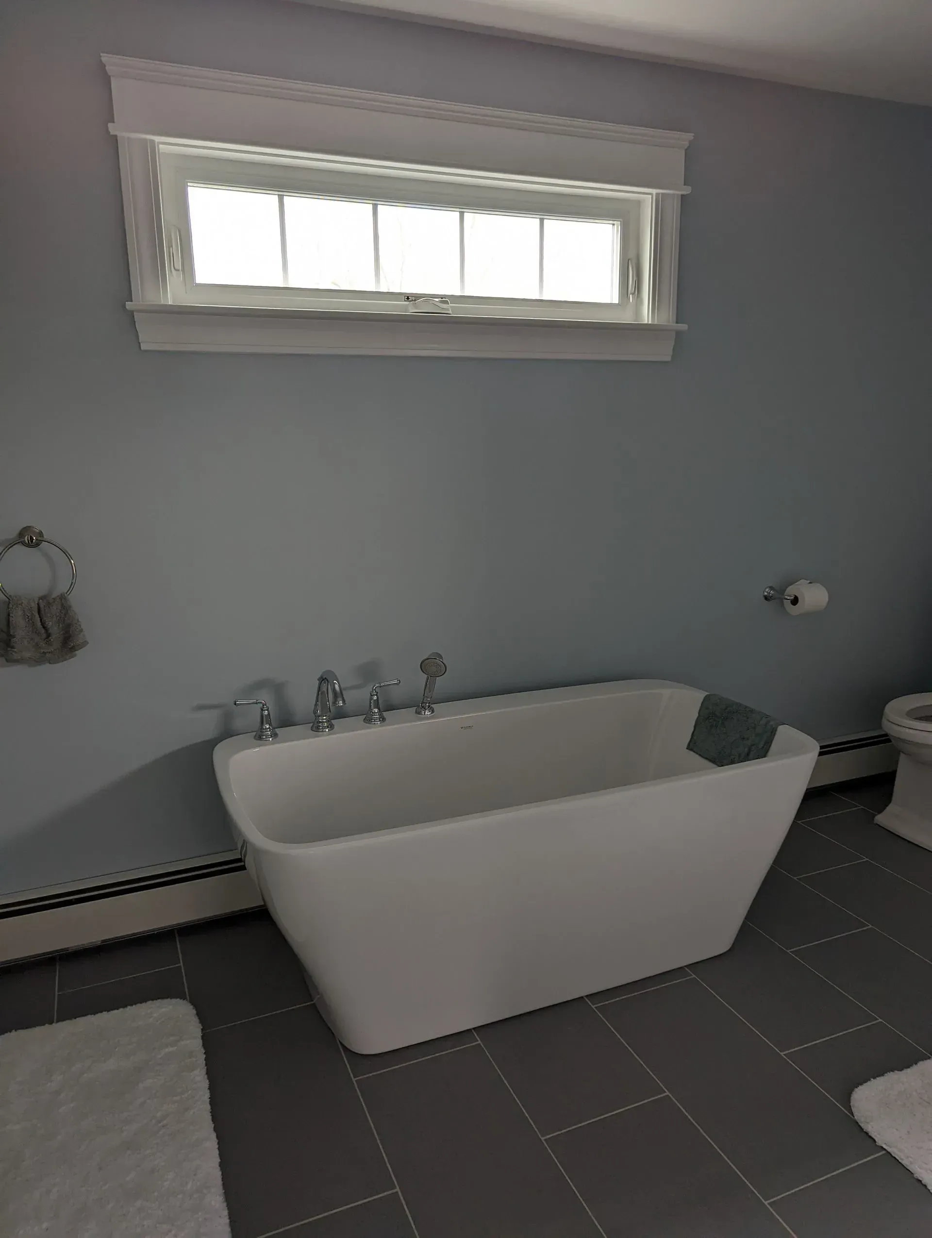 Bright bathroom with freestanding white tub, gray walls, and a small window above it.