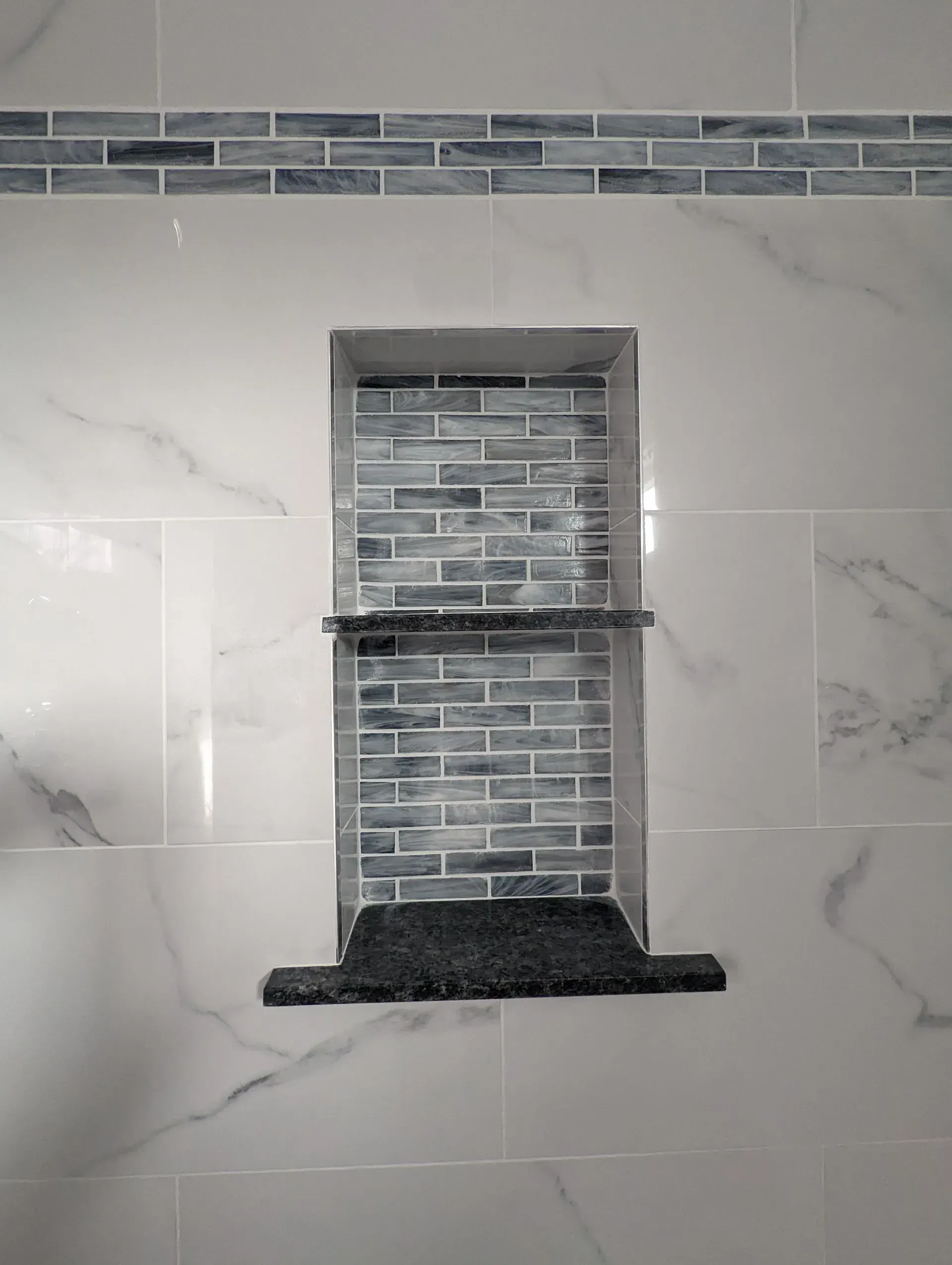 Shower wall with recessed tiled niche and dark stone shelf, surrounded by white marble-look tile.