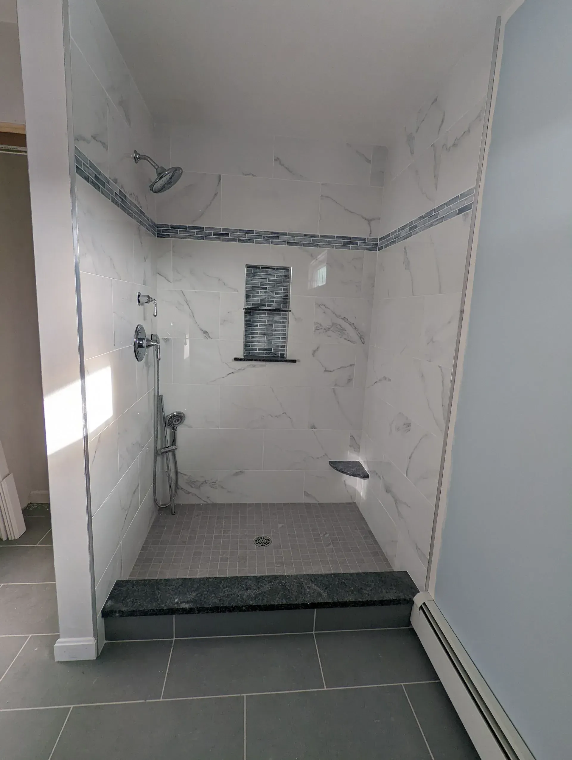 Small tiled shower stall with white marble walls, gray floor, and a chrome handheld shower.
