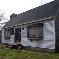Small white house with dark roof, open front door, and windows with shutters.