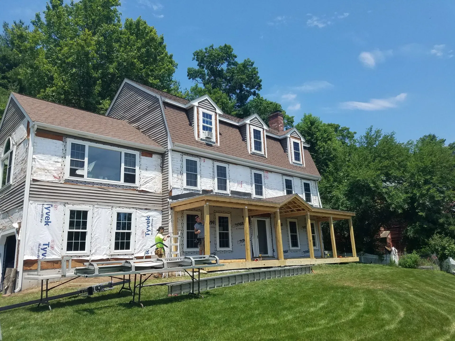 Large white farmhouse with yellow porch and dormer windows on a green lawn under a blue sky.