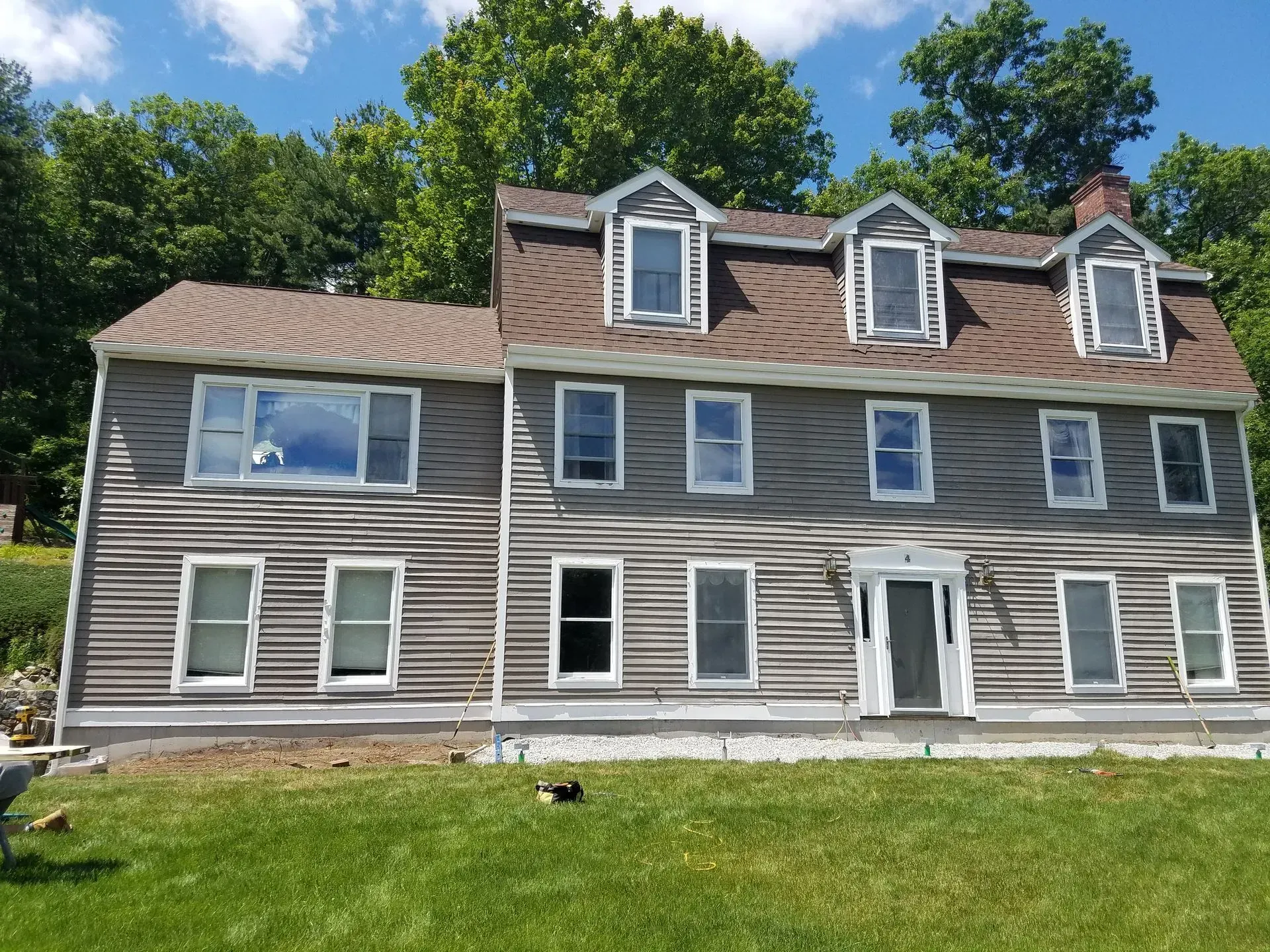 Gray two-story house with white trim and dormer windows on a grassy hill under a blue sky