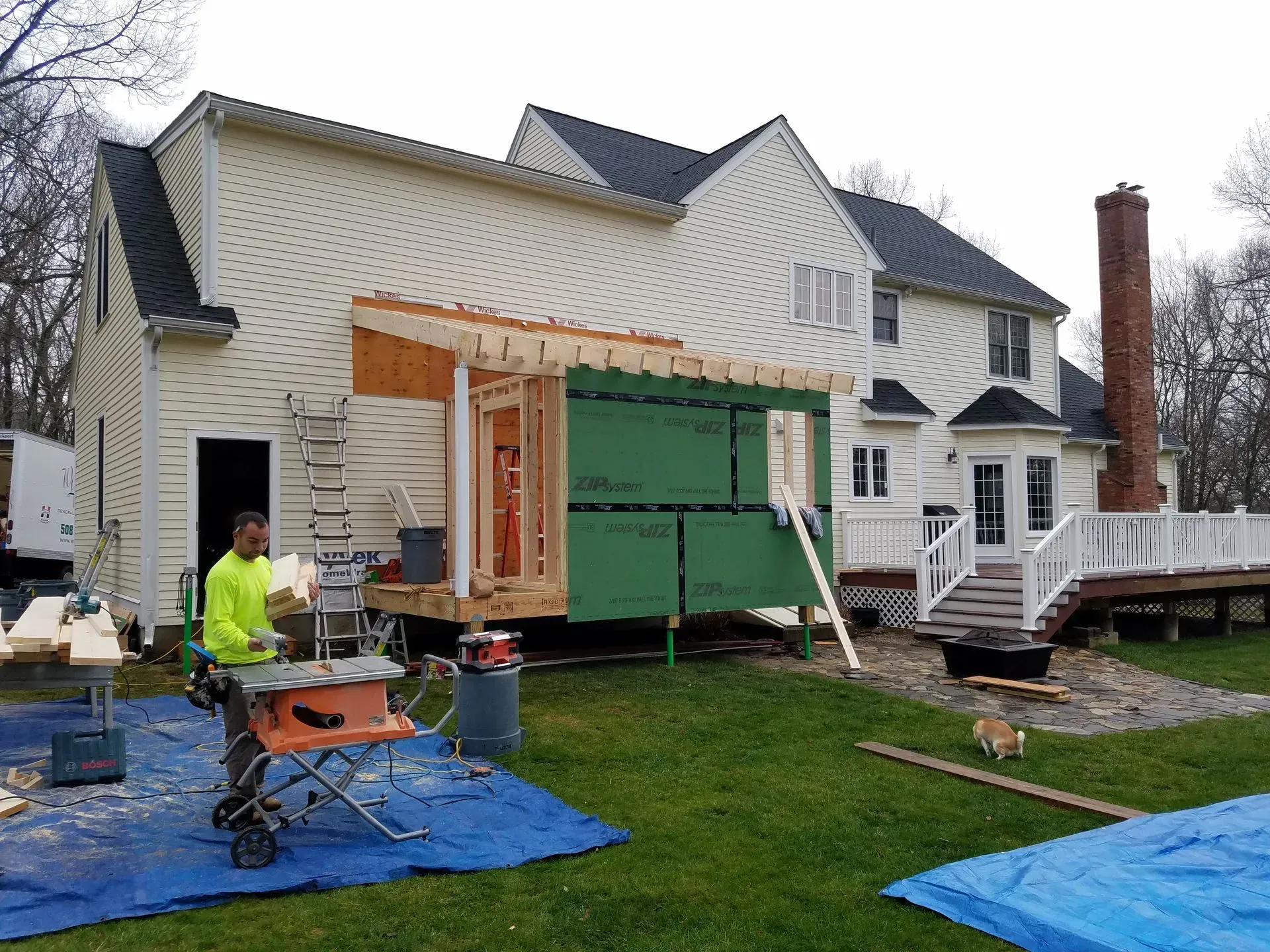 Workers building a house addition with green sheathing and exposed framing beside a white home