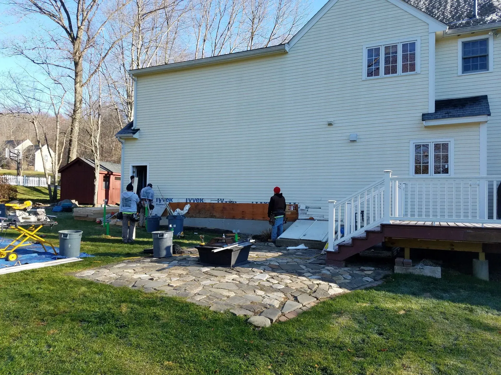 Backyard patio beside a white house, with two people standing near a stone fire pit and a raised deck.