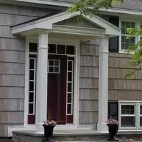 Front entrance of a gray house with white columns and a red door, flanked by potted flowers