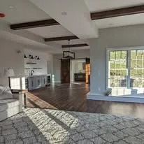 Bright open living room with white kitchen, dark wood floors, exposed beams, and large windows.