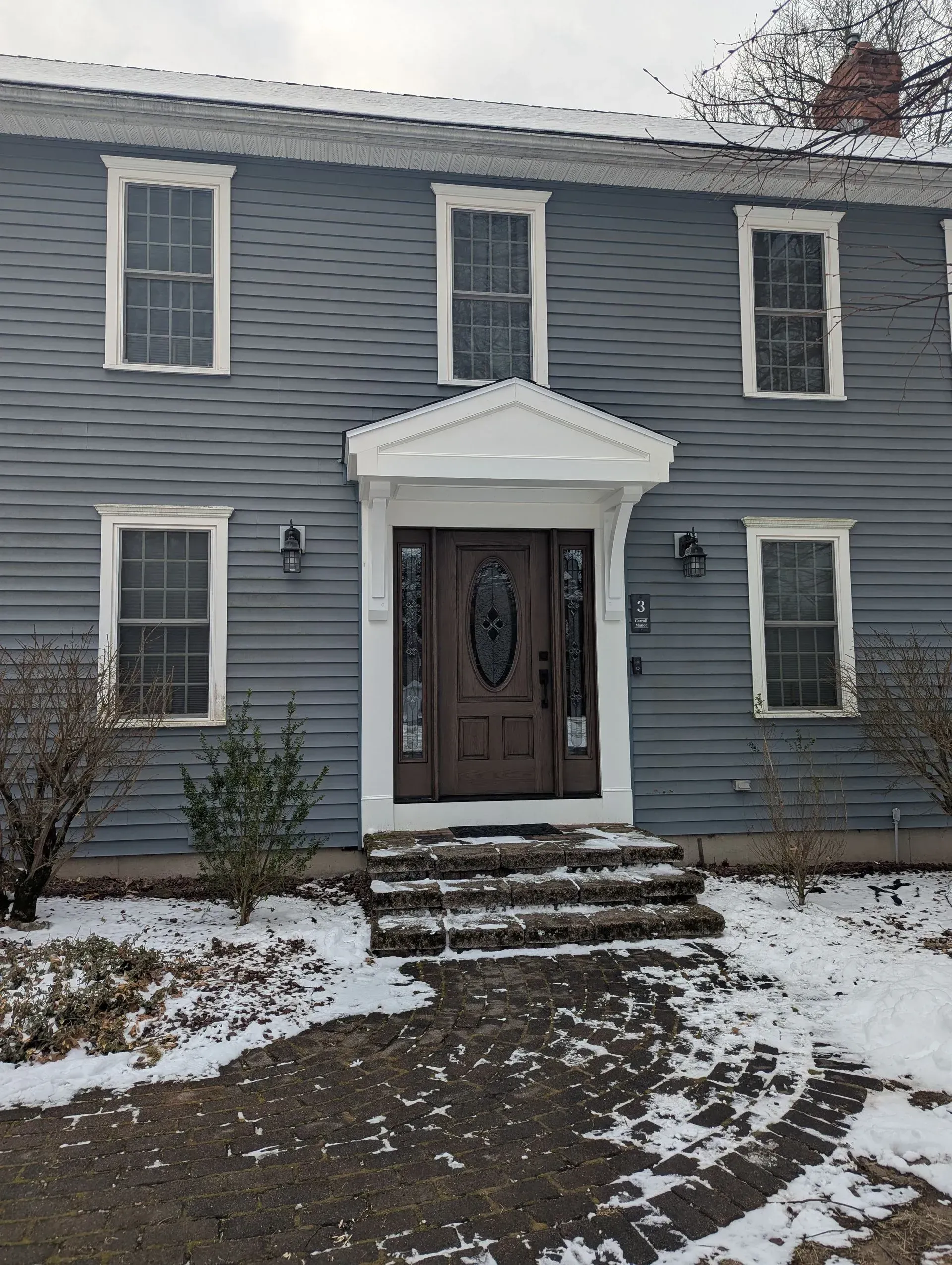 Blue two-story house with white trim, snow on the ground, and a brown front door with steps leading up to it