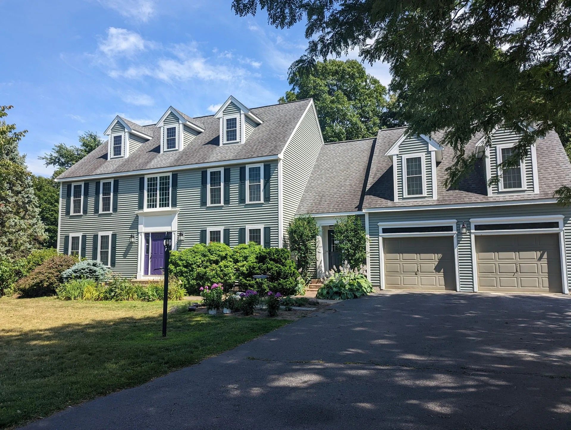 Large green house with white trim and double garage, viewed from a driveway under trees