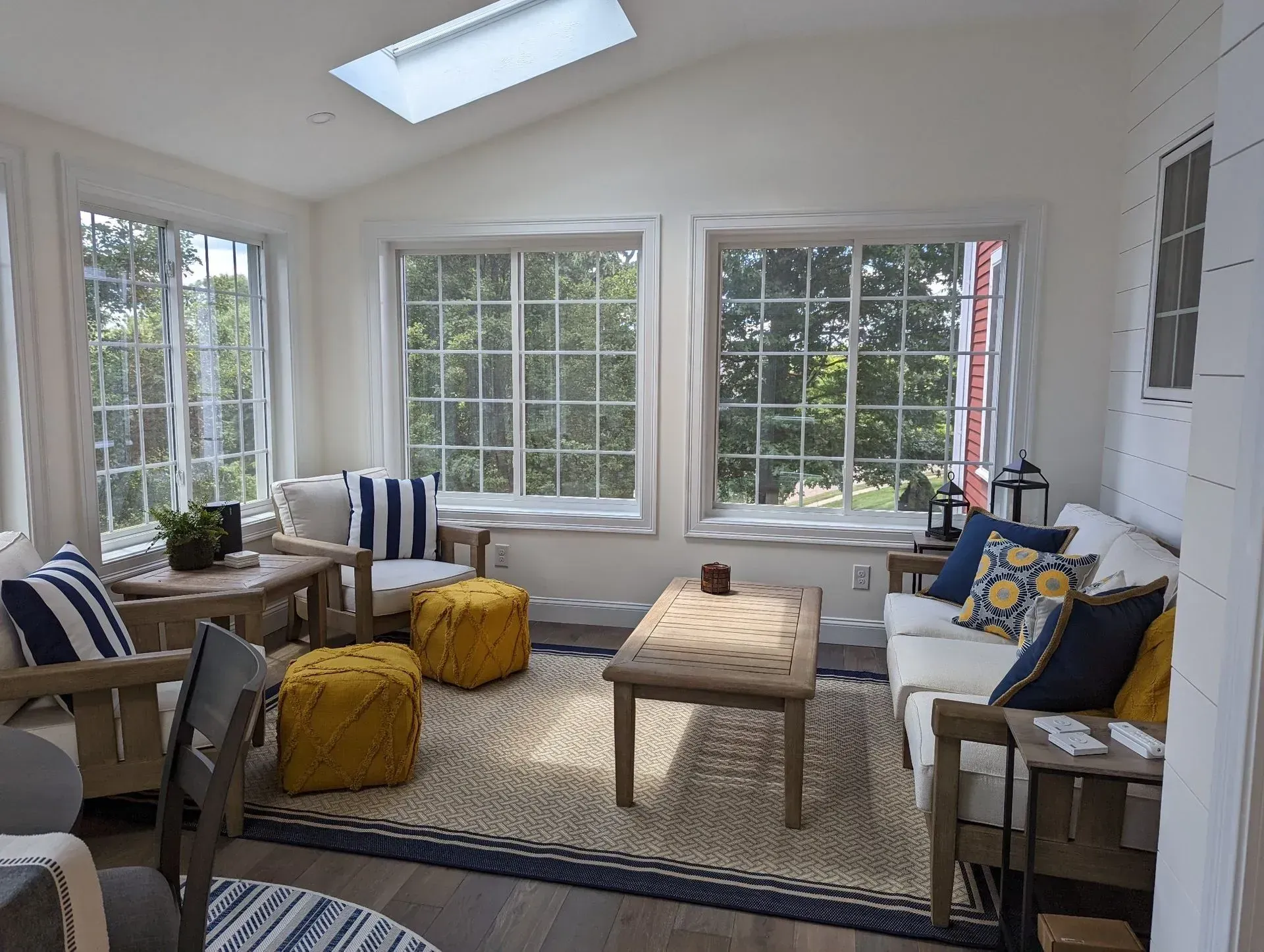 Bright sunroom with white walls, large windows, wicker chairs, and a beige ottoman on a patterned rug.