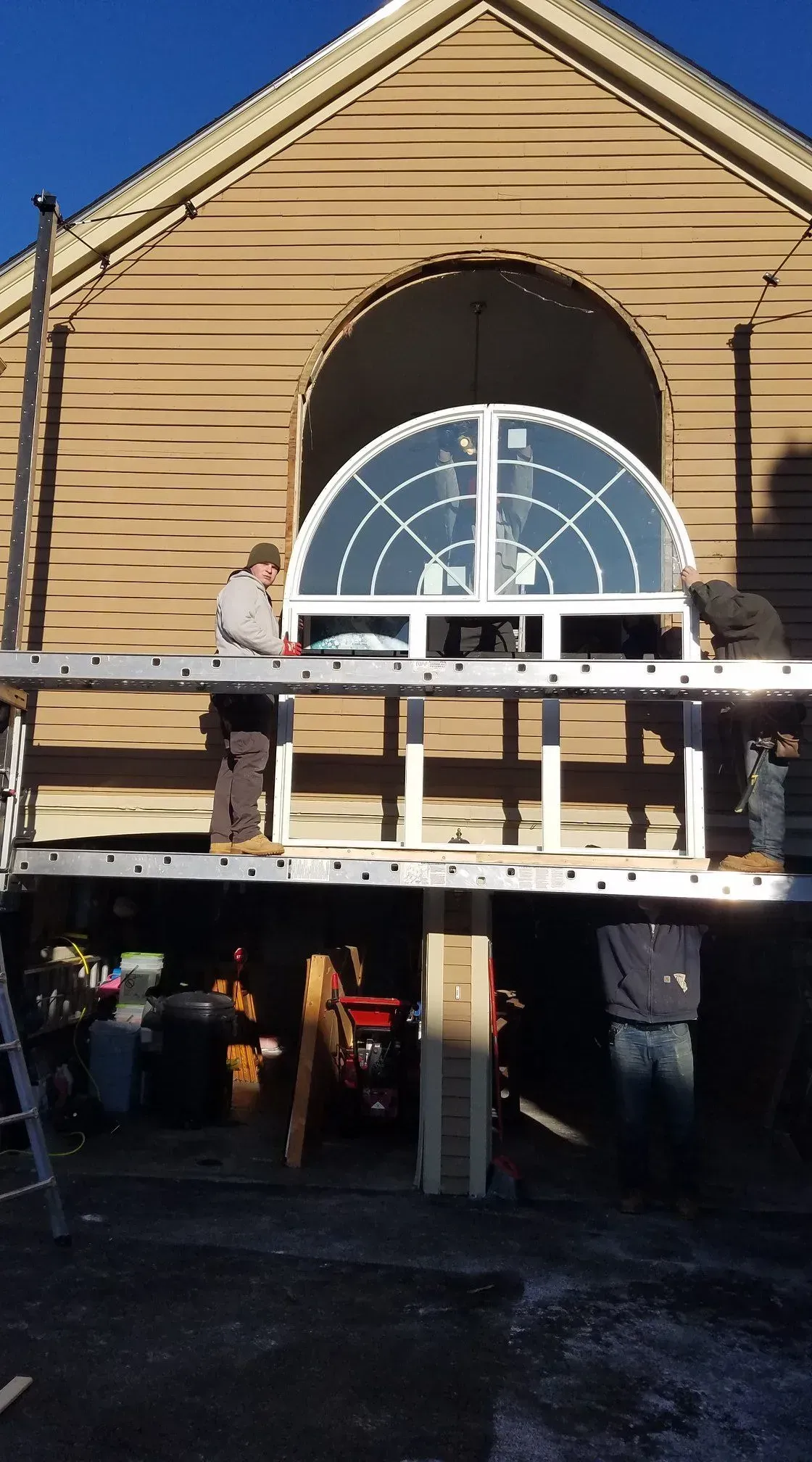 Two people on a scaffold installing a large arched window on a tan building exterior