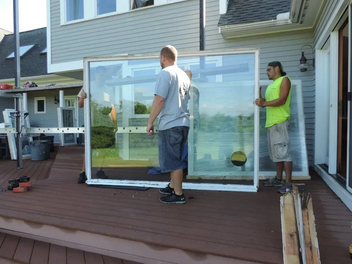 Two workers carry a large glass pane onto a wooden deck beside a house