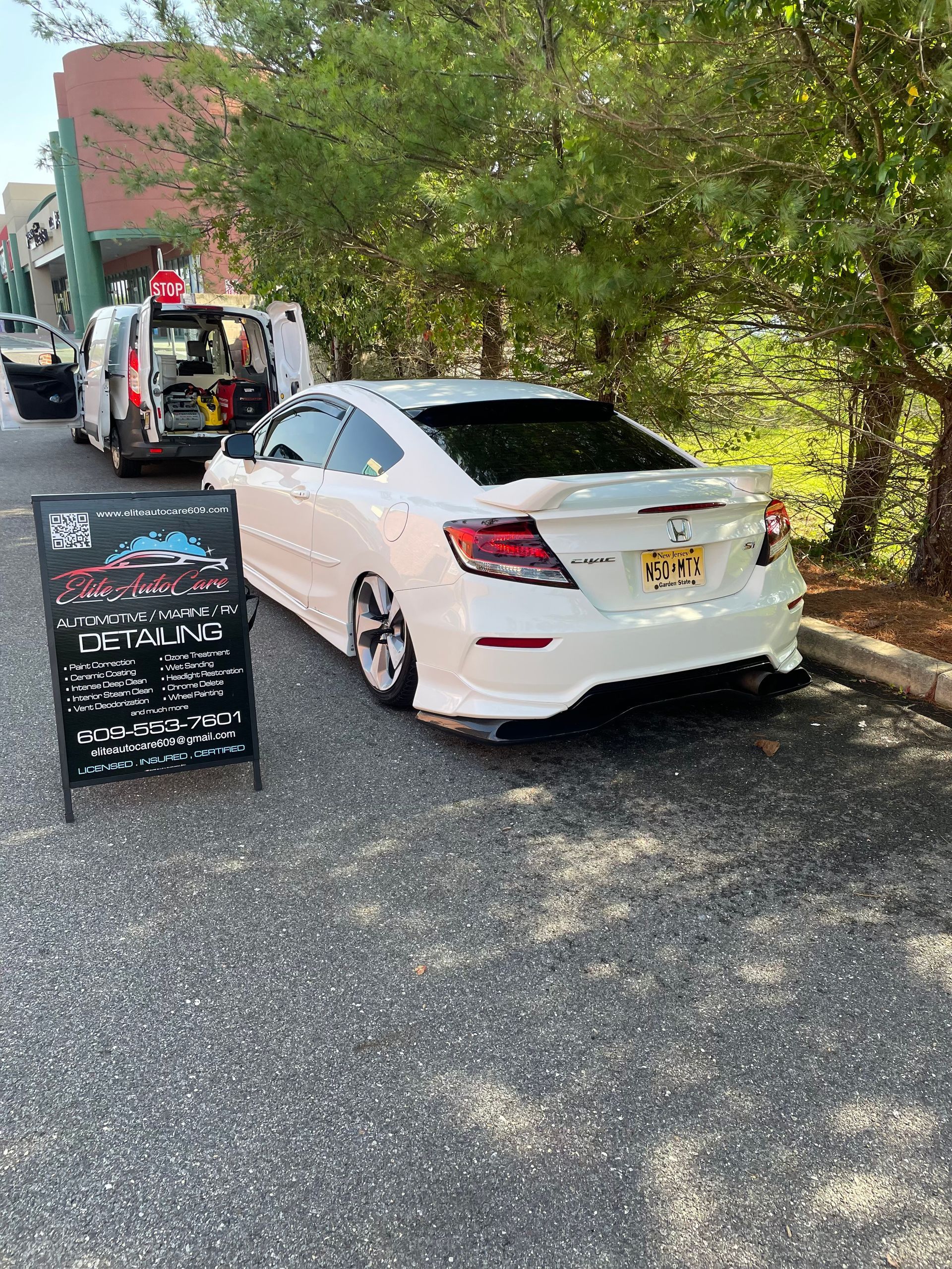 White sedan parked on gravel beside a chalkboard sign and trees at an outdoor lot