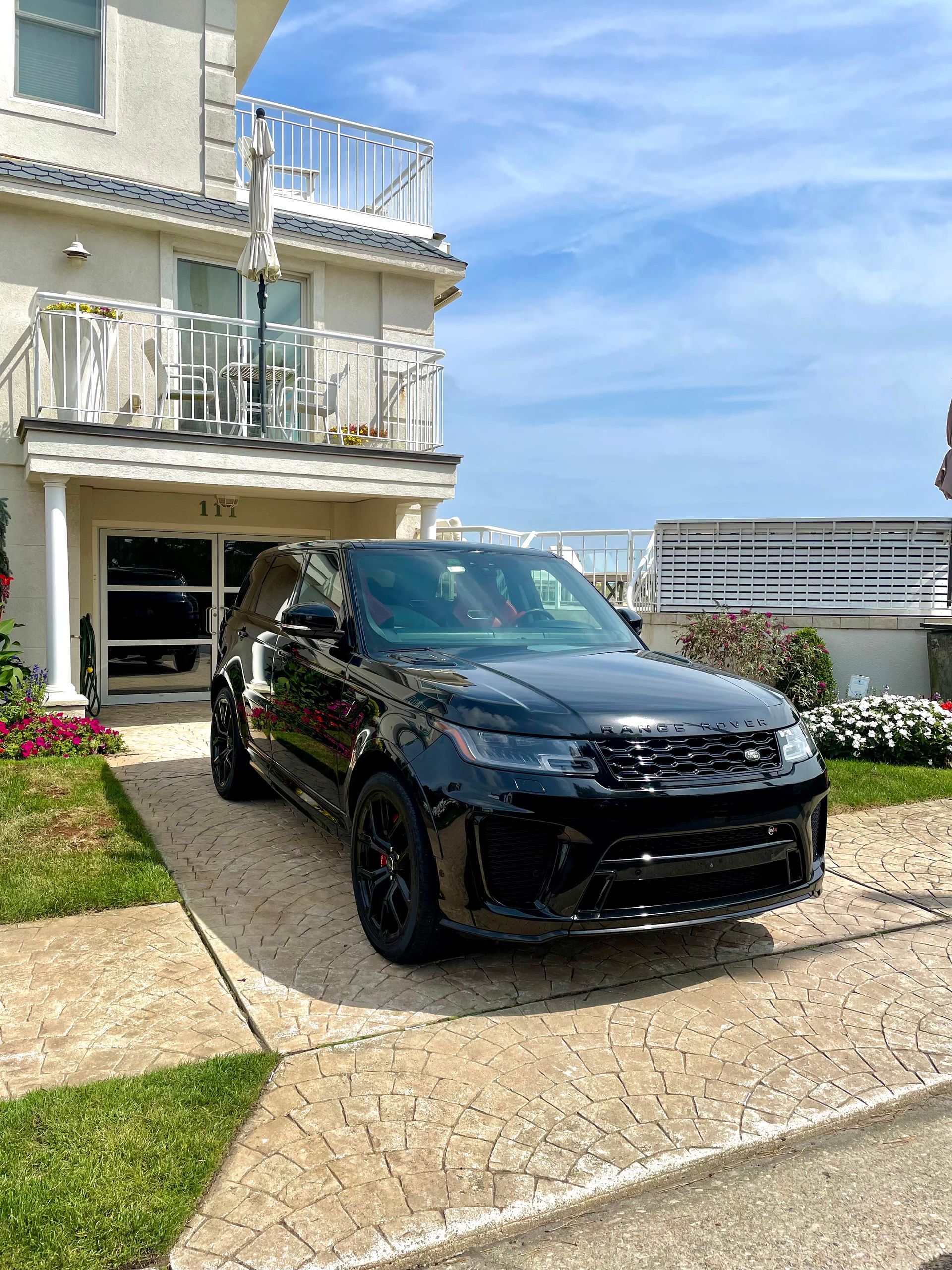 Black SUV parked in a driveway beside a beige house with balconies on a sunny day