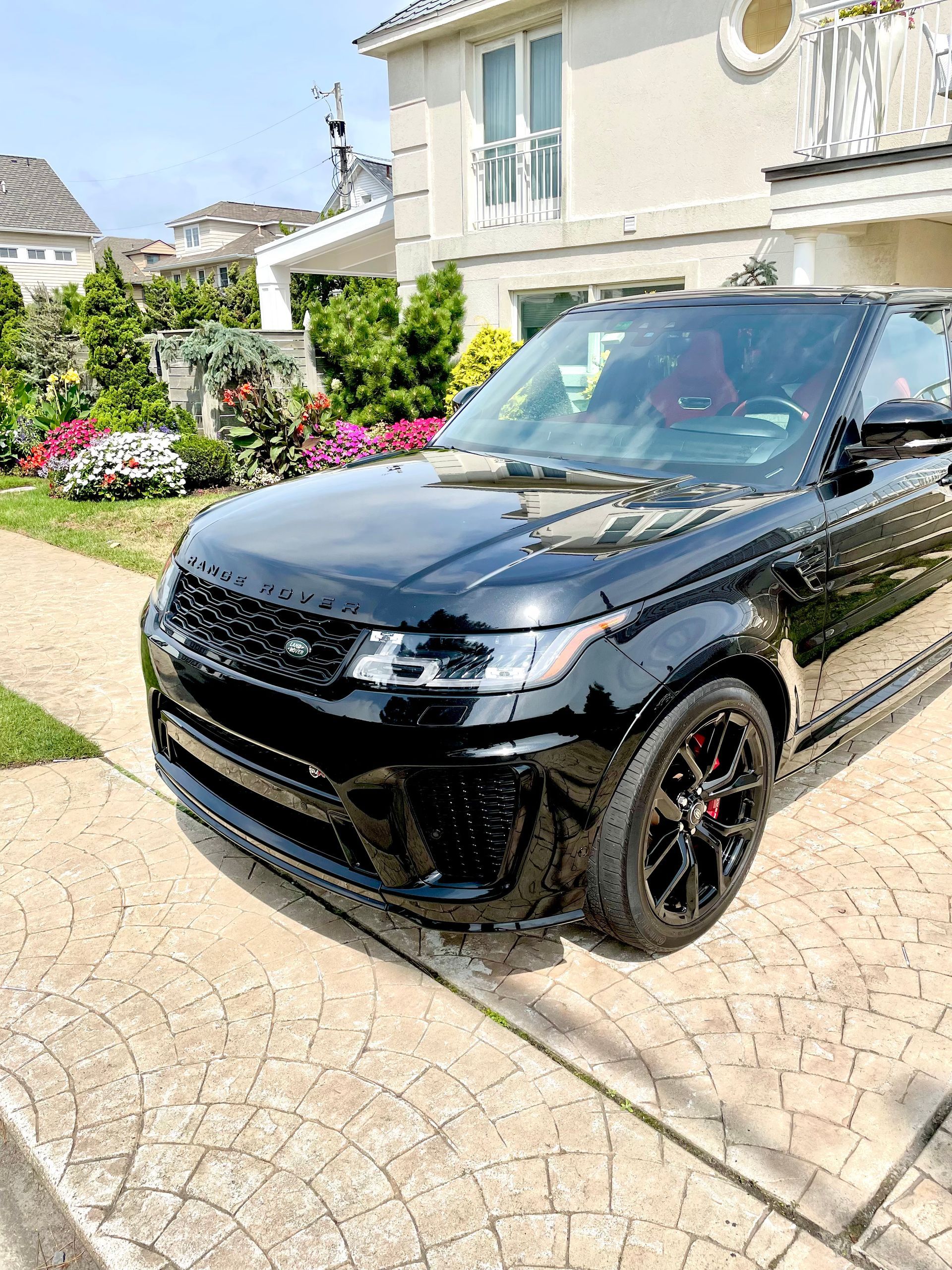 Black SUV parked in a sunny driveway beside a beige house with flowers and greenery.