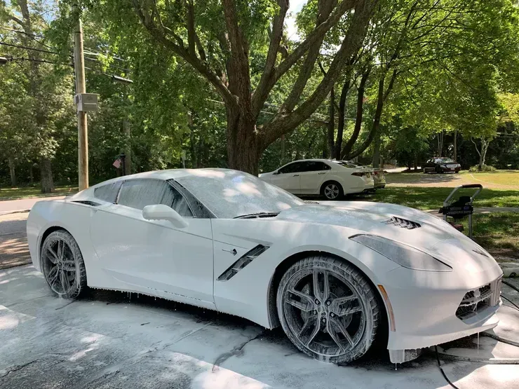 White sports car parked on a snowy street under trees, with another car in the background.