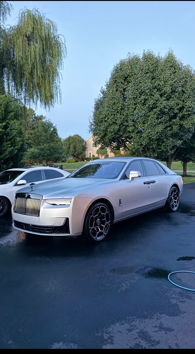 Silver luxury sedan parked on a wet driveway beside a white car, with trees and a house in the background.